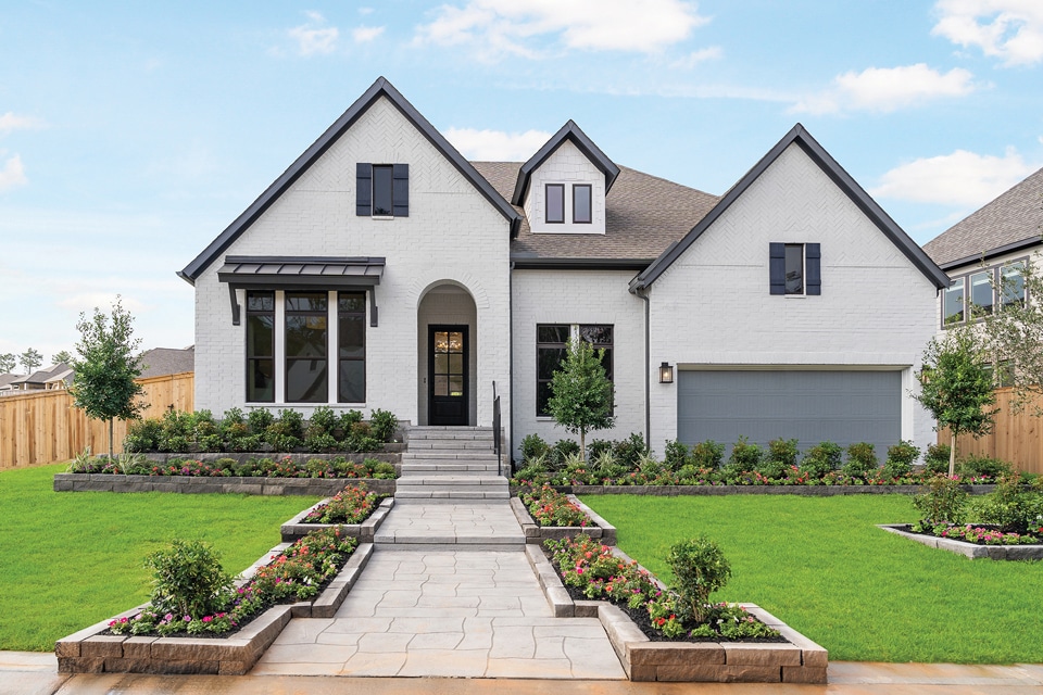 A modern two-story house in Medina Valley with a white brick exterior, black trim, a two-car garage, manicured lawn, and landscaped flower beds along a stone walkway—perfect for new homes near San Antonio.