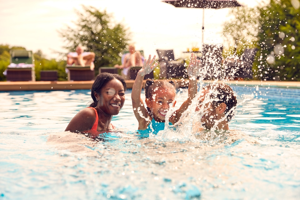 Three people play and splash water in an outdoor swimming pool on a sunny day at Briggs Ranch, a master-planned community in the scenic Medina Valley, with others relaxing in the background.