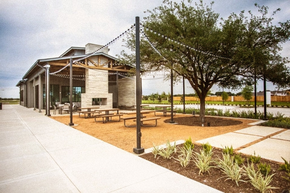 Outdoor patio area with picnic tables, string lights, and a large tree set beside a modern building under a cloudy sky—perfect for relaxing at new homes in Briggs Ranch, San Antonio.
