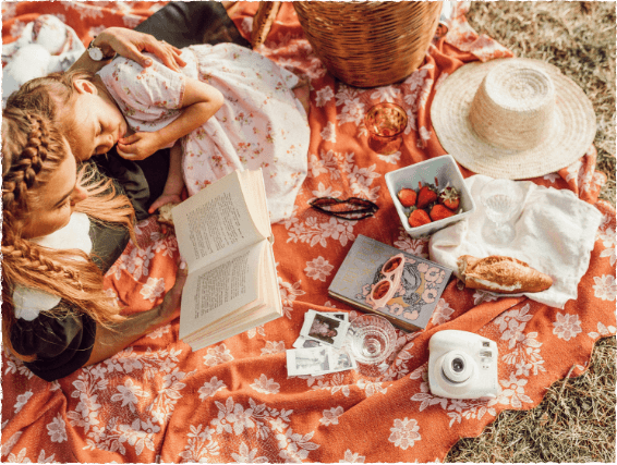 Woman and child lying on a floral blanket having a picnic with strawberries, bread, a hat, book, sunglasses, basket, instant photos, and an instant camera—enjoying sunny days in Medina Valley near new homes in San Antonio.