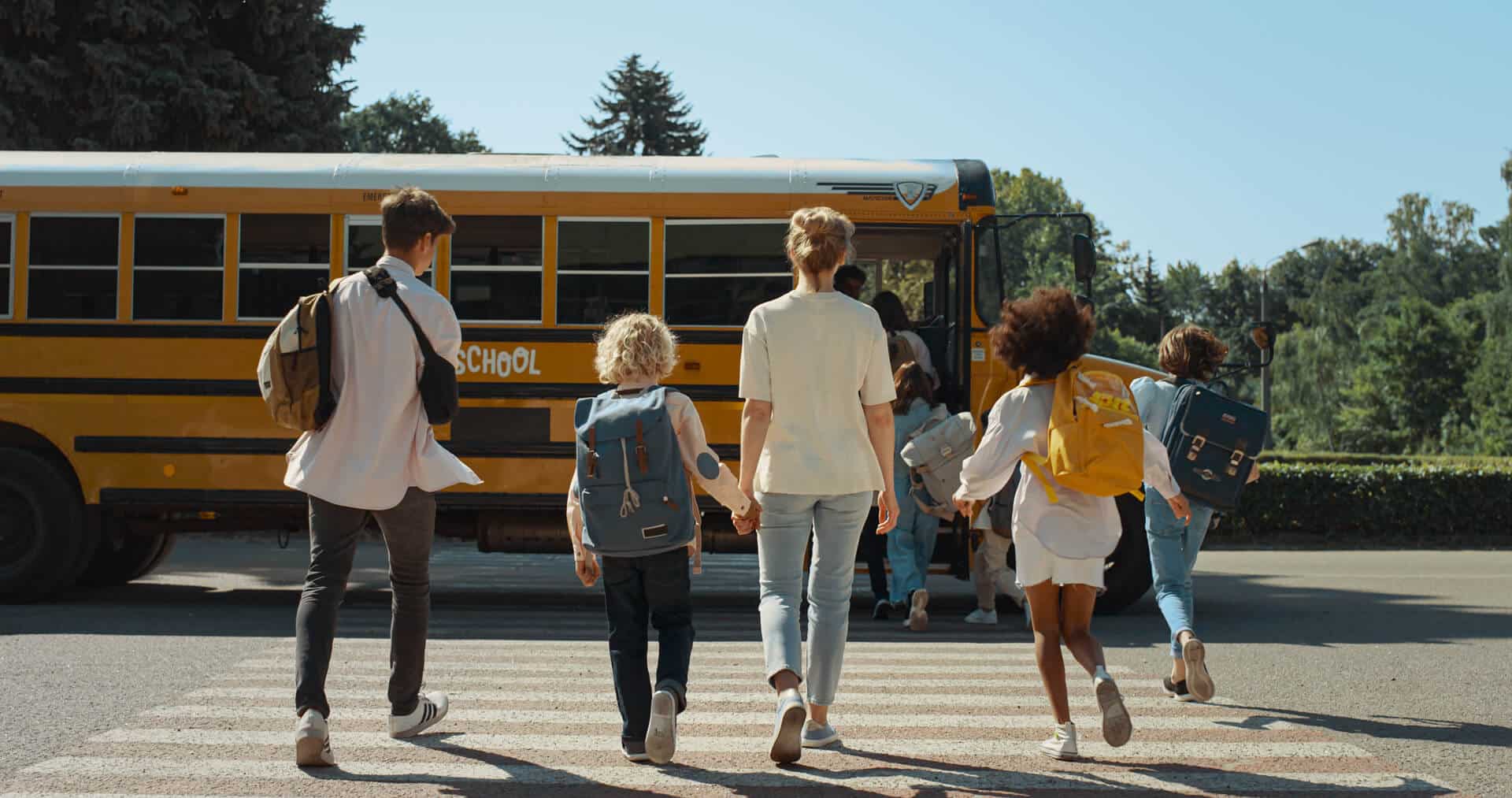 A group of children and an adult cross a street toward a yellow school bus on a sunny day in Medina Valley, carrying backpacks near the new homes of Briggs Ranch.
