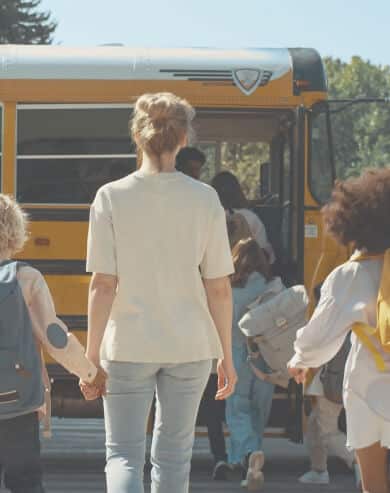An adult and several children walk toward a yellow school bus in a master-planned community near Medina Valley, San Antonio, while children board through the open door.