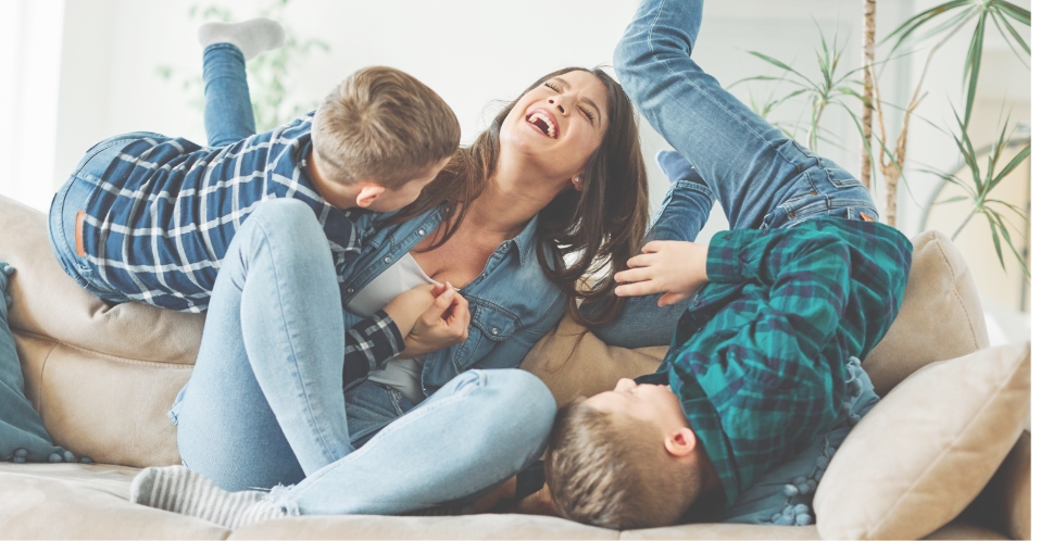 A woman sits on a couch laughing while two children in plaid shirts and jeans playfully climb around her, enjoying family time in their new home within the vibrant Medina Valley master-planned community.