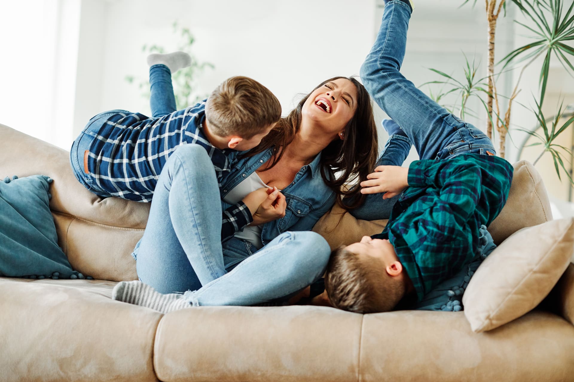 A woman sitting on a couch laughs while two children in plaid shirts play energetically around her, capturing the joyful spirit of family life in a master-planned community like Medina Valley with its inviting new homes.