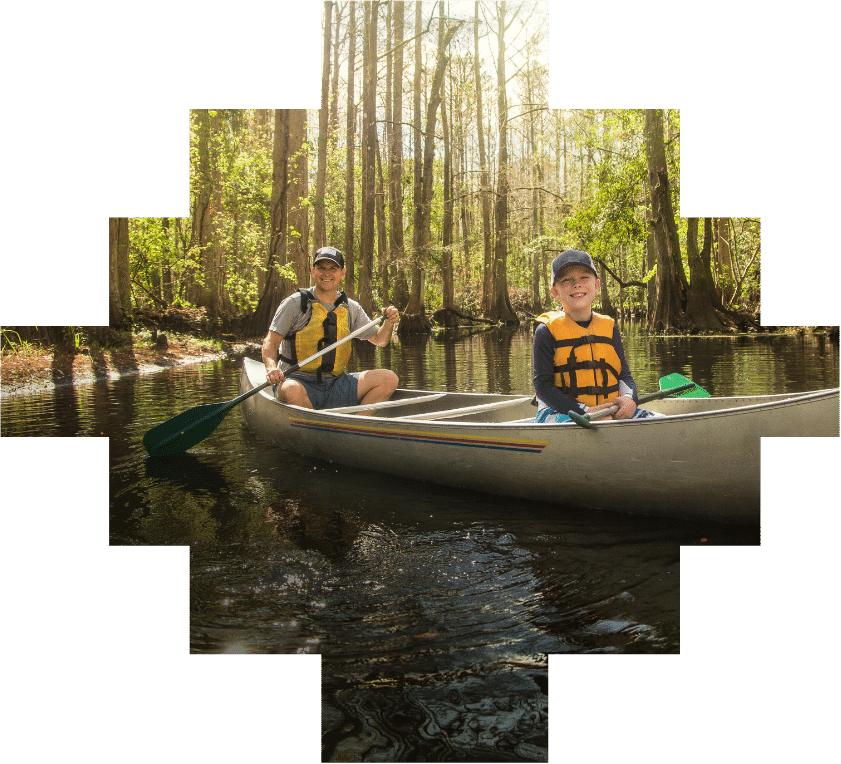 An adult and a child wearing life jackets paddle a canoe on a calm, wooded river near Briggs Ranch in San Antonio, exploring nature just minutes from new homes for sale.
