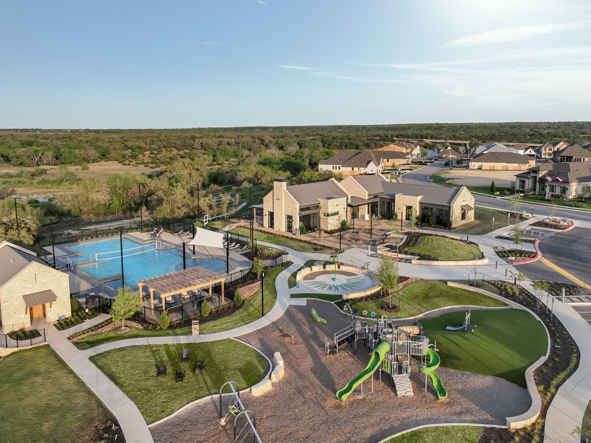 Aerial view of a suburban community park in San Antonio’s Briggs Ranch, featuring a playground, splash pad, swimming pool, tennis court, clubhouse buildings, and landscaped walkways near beautiful new homes.