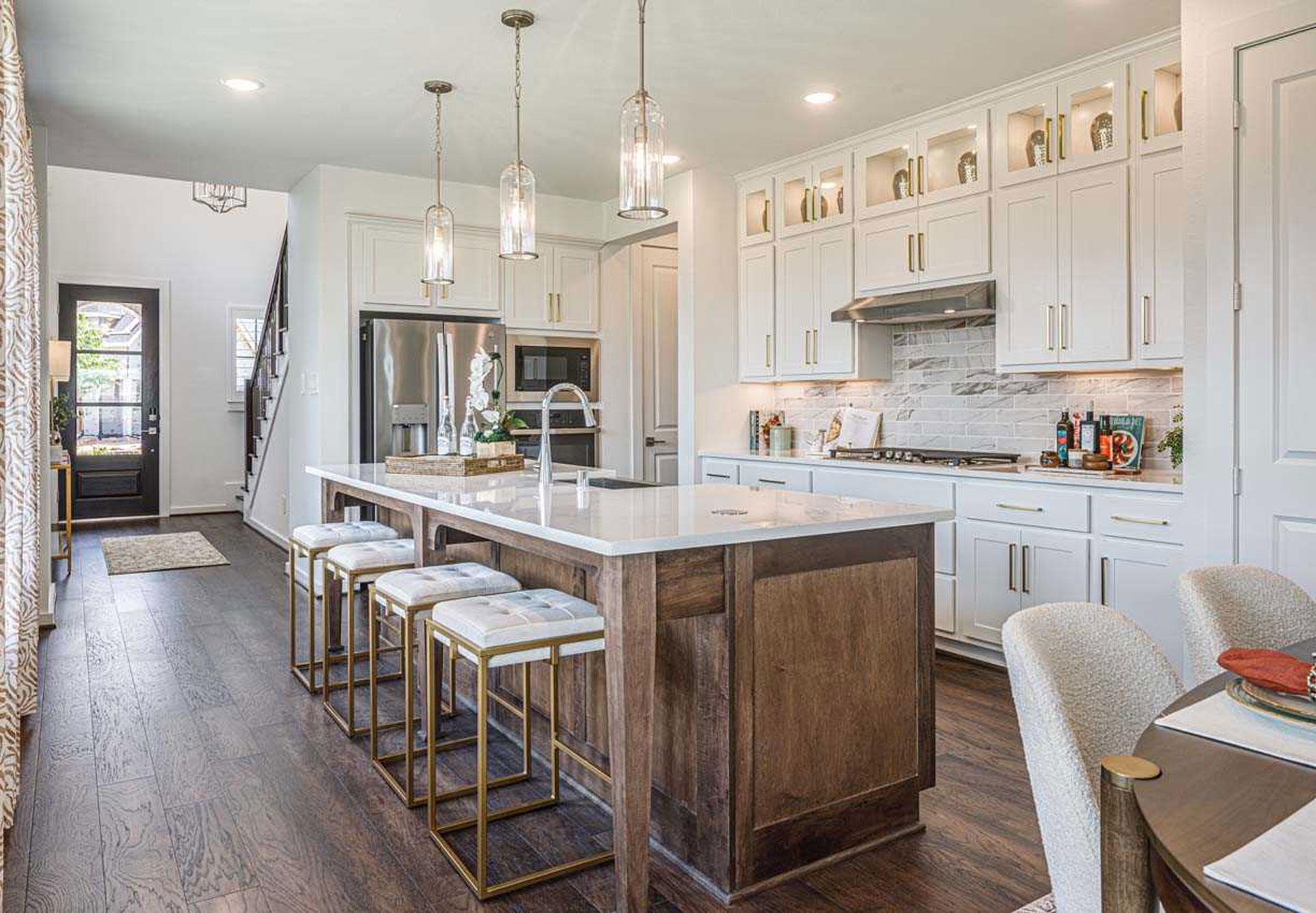 Modern kitchen in San Antonio with white cabinets, a large island with wood accents, three stools, pendant lights, stainless steel appliances, and a dining table—perfect for those seeking new homes for sale in the Medina Valley area.