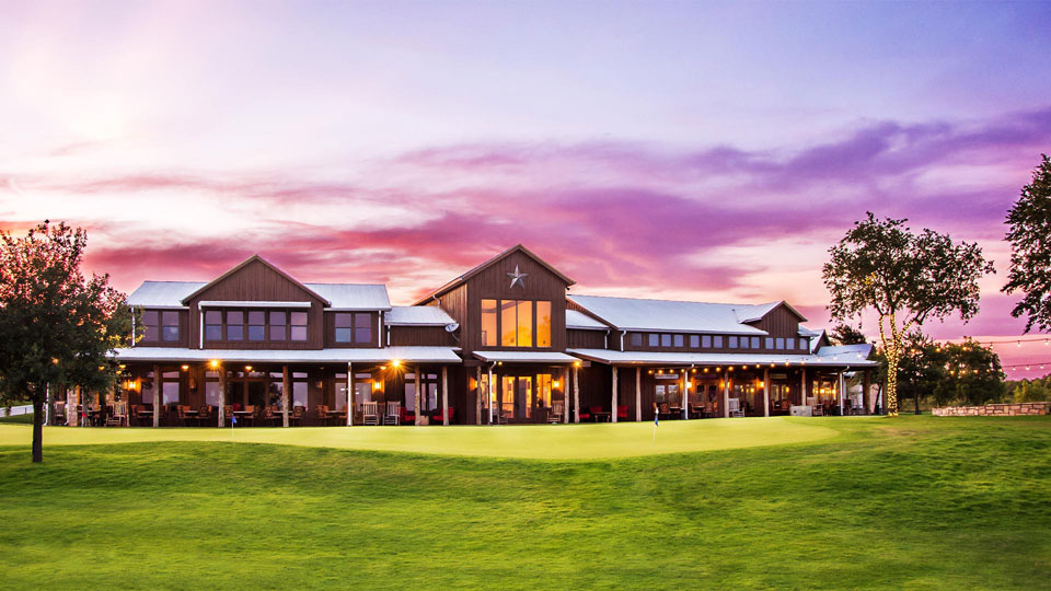 A large modern ranch-style building with wide windows, outdoor seating, and a manicured lawn, set against a colorful sunset sky—perfectly located in the heart of Medina Valley’s master-planned community with new homes for sale.