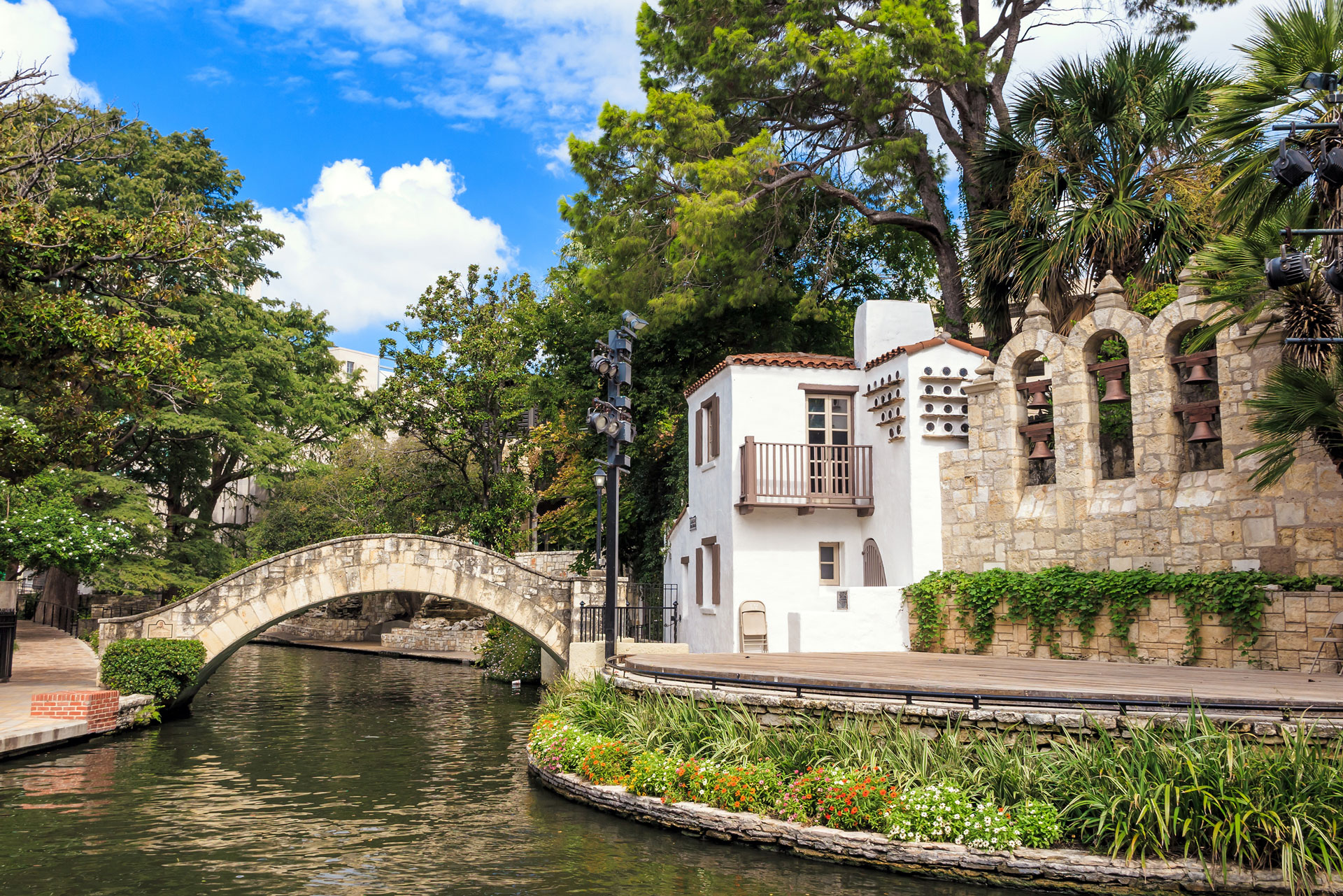A stone footbridge arches over a narrow river beside a white stucco building with a balcony, surrounded by greenery and trees under a blue sky—capturing the charm of San Antonio near beautiful new homes for sale.