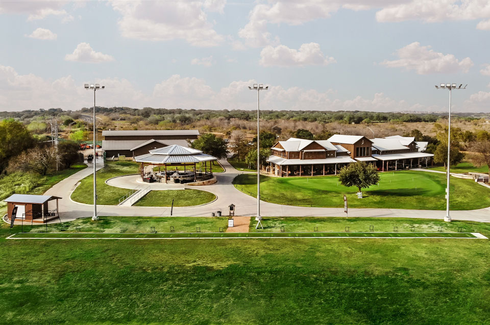 An aerial view of Briggs Ranch showcases a large green lawn with a covered pavilion, new homes for sale, winding pathways, trees, and light poles under a partly cloudy Medina Valley sky.