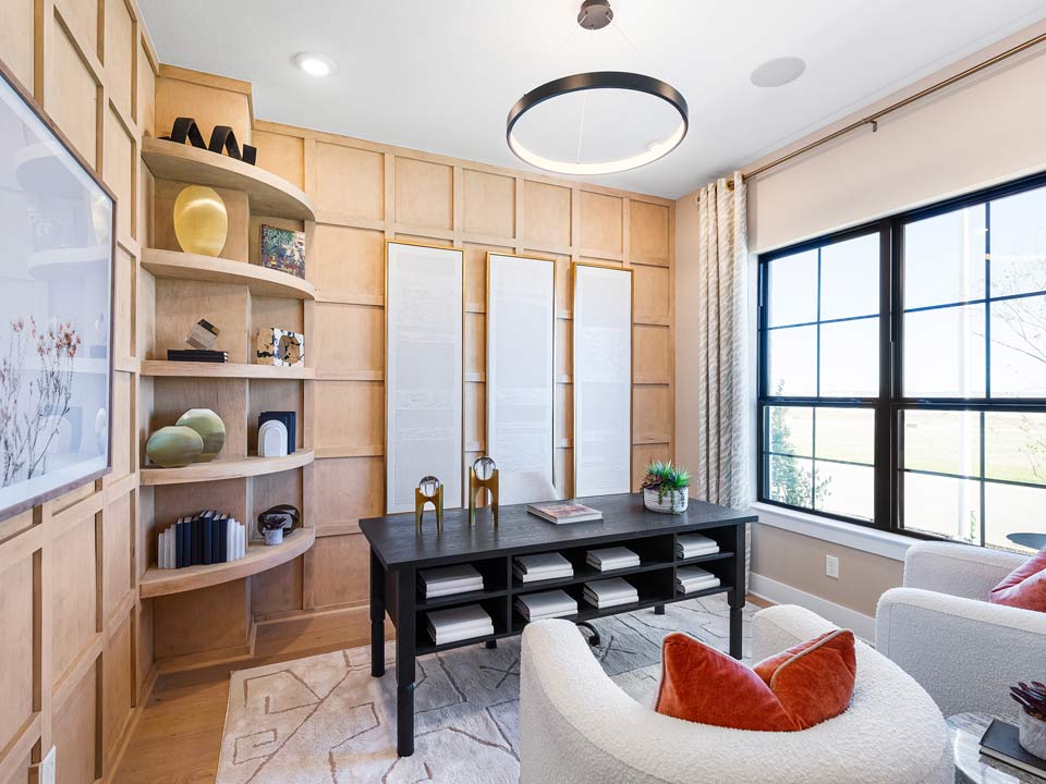 Modern home office with light wood paneled walls, black desk, white armchairs with orange pillows, open shelves, and a large window letting in natural light—perfect for new homes in a master-planned community near San Antonio.