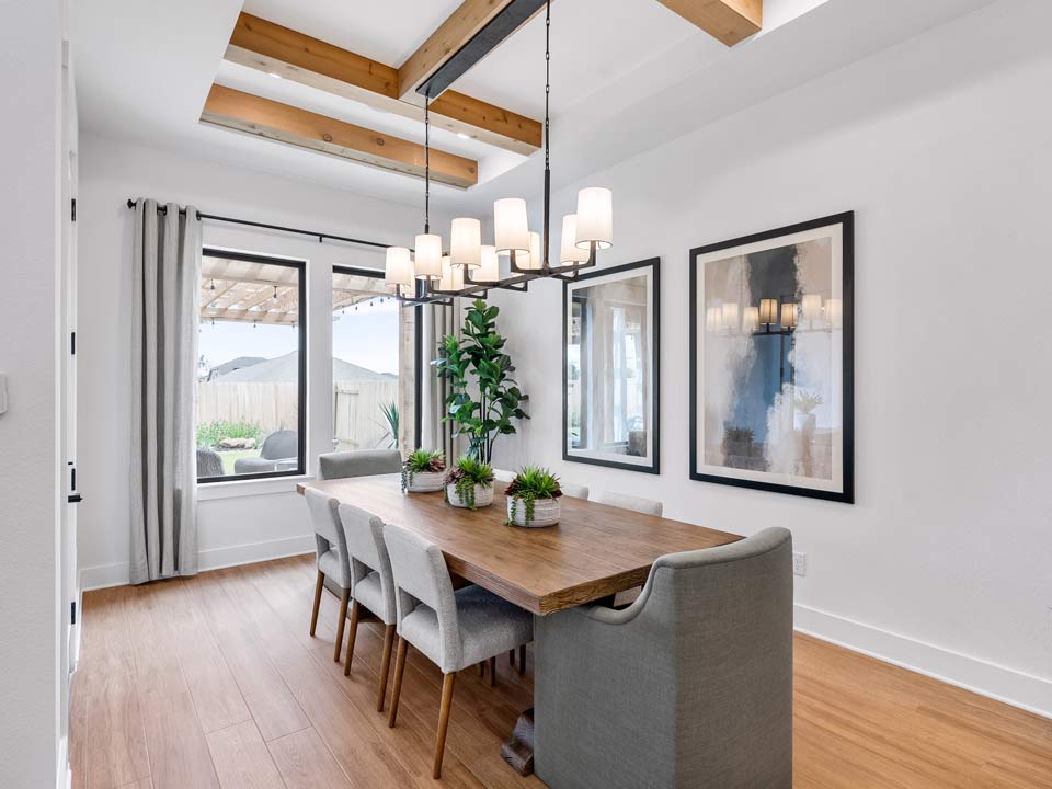 Modern dining room in a master-planned community, featuring a wooden table, gray upholstered chairs, chandelier, potted plants, and framed wall art. Large windows and wooden ceiling beams bring warmth—perfect for San Antonio living.