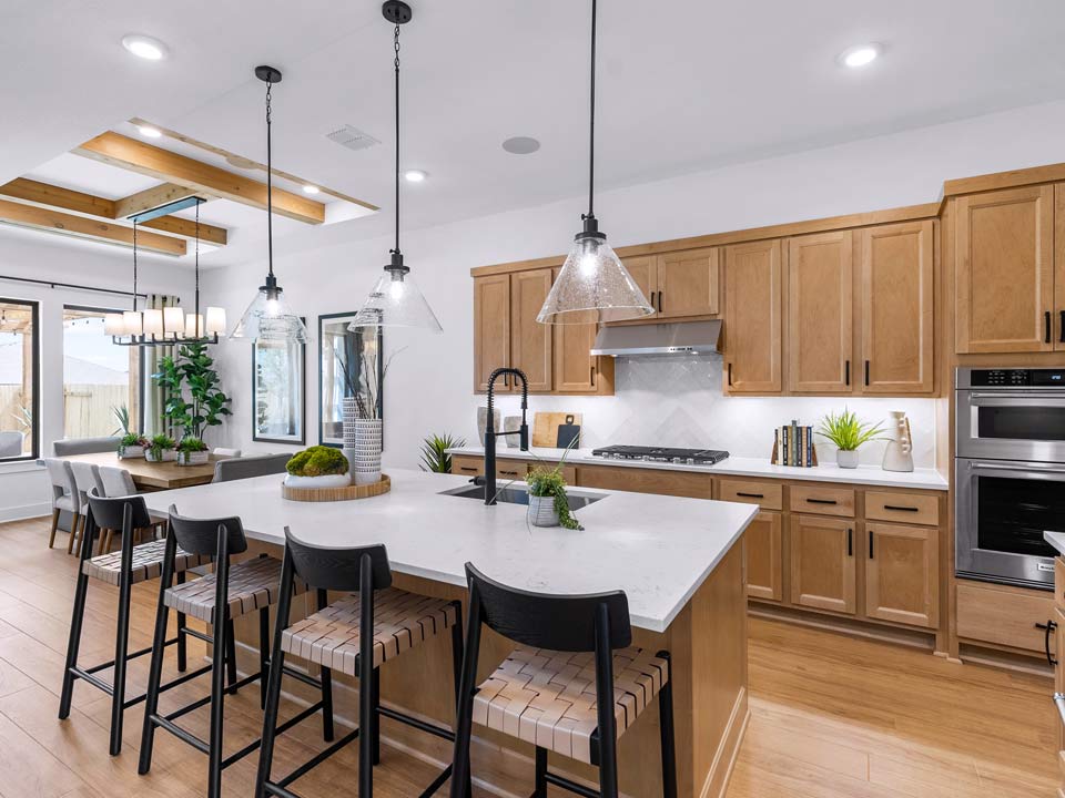 Modern kitchen with wooden cabinets, white countertops, a large island with four chairs, pendant lighting, and stainless steel appliances. This stunning space is featured in new homes for sale in Medina Valley near San Antonio. Dining area visible in the background.
