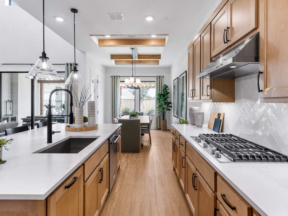 Modern kitchen with light wood cabinets, white countertops, stainless steel appliances, pendant lights, and a view into a dining area with large windows in a San Antonio master-planned community.