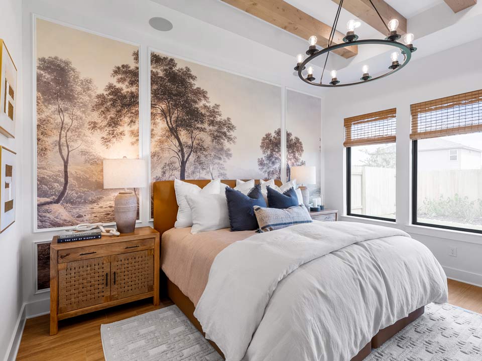 A cozy bedroom in Medina Valley, featuring a large bed, neutral bedding, rustic nightstand with lamp, a wall mural of trees, wooden ceiling beams, and windows with bamboo shades—perfect for those seeking new homes for sale near San Antonio.