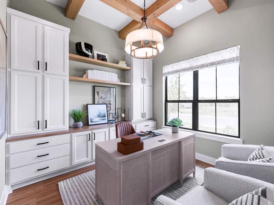 A modern home office in a master-planned community features a wooden desk, white cabinets, wall shelves, two armchairs, a pendant light, and a large window welcoming natural light—perfect for new homes in San Antonio.
