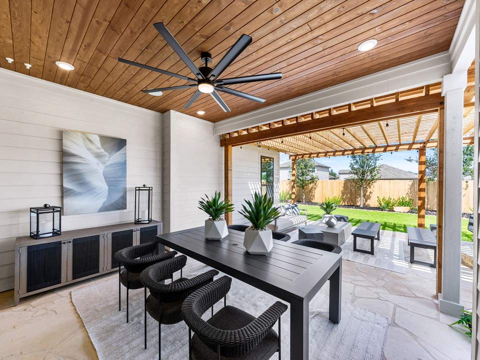 Modern covered patio in San Antonio featuring a black dining table with four wicker chairs, potted plants, ceiling fan, artwork, and a view of the backyard pergola—perfect for relaxing or entertaining in new homes for sale in Medina Valley.