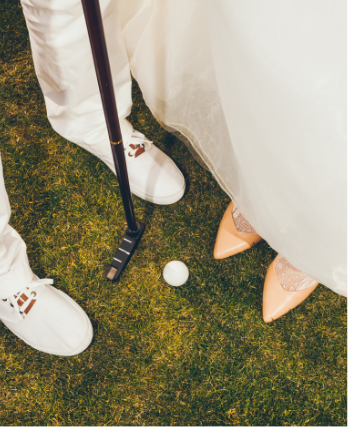 Two people stand on grass in Medina Valley, one holding a golf club near a golf ball. One wears white shoes; the other wears nude heels and a white dress—a perfect day in San Antonio, where new homes for sale are nearby.