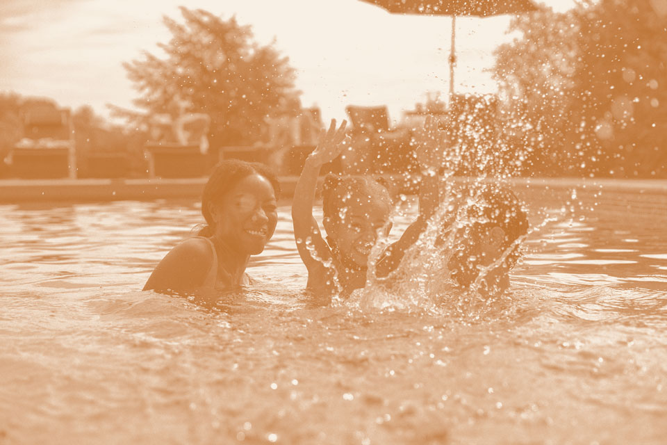 Three children are playing and splashing water in a swimming pool outdoors, with sunlight illuminating the scene in a vibrant new homes community near Medina Valley, San Antonio.