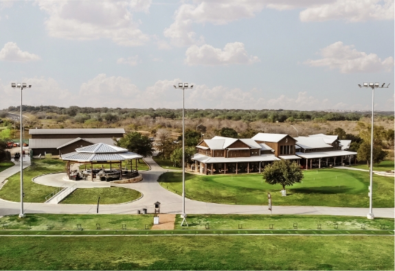 Aerial view of a recreational facility at Briggs Ranch in San Antonio, featuring a large building, pavilion with picnic tables, green lawns, and tall light poles under a partly cloudy sky—perfect for enjoying new homes nearby.