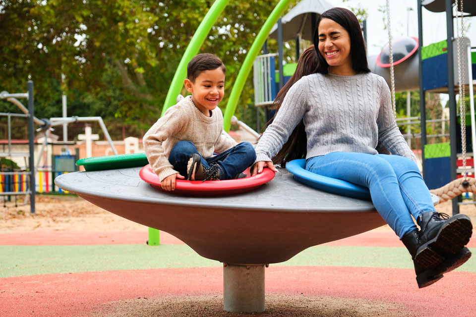 A woman and a young boy sit on a spinning playground apparatus at Briggs Ranch, smiling and holding hands, with playground equipment visible in the background—perfect family fun near new homes for sale in San Antonio.