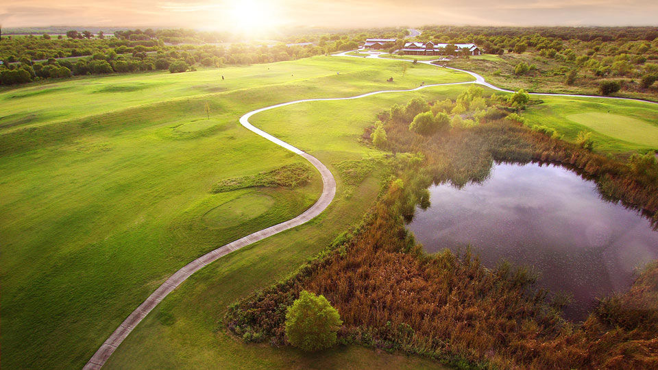 Aerial view of a golf course at sunset in Medina Valley, with green fairways, a winding path, a pond, scattered trees, and new homes for sale visible near the distant buildings.