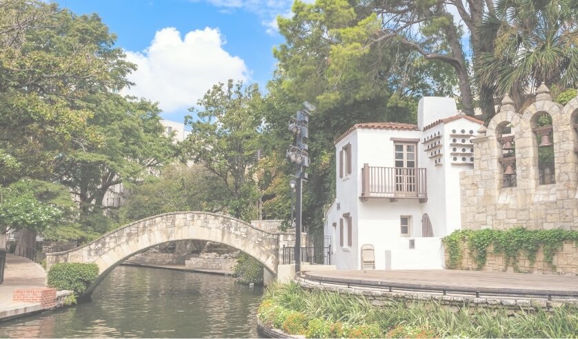 A stone footbridge arches over a river near a white stucco building and lush green trees under a partly cloudy sky, capturing the tranquil charm of Briggs Ranch in San Antonio, where new homes for sale await.