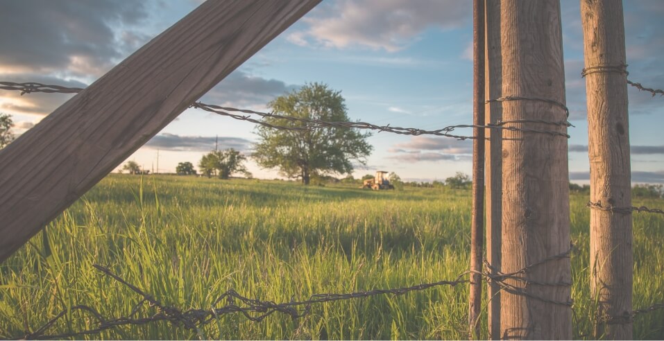 Barbed wire fence in the foreground with a grassy field, a tree, and a tractor in the distance under a partly cloudy sky at sunset, capturing the rural charm of Medina Valley near San Antonio—perfect for those seeking new homes for sale.