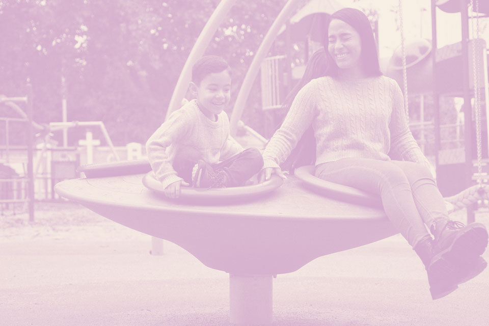 A woman and a child sit and smile together on a circular playground spinner at a park in a master-planned community, enjoying quality time surrounded by new homes for sale.
