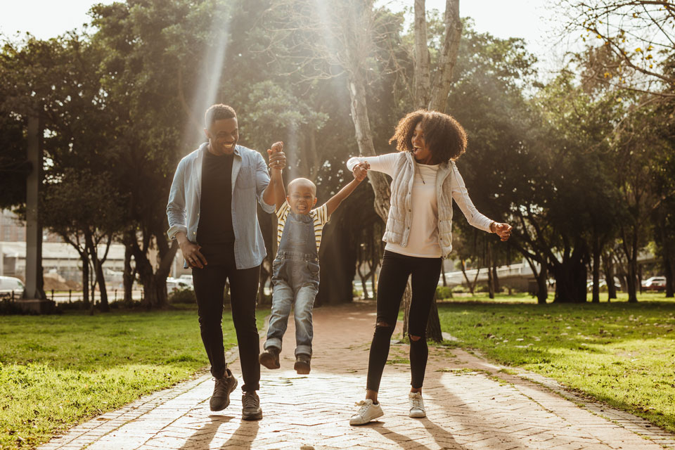 Two adults swing a smiling child between them as they walk along a sunlit path in a park, surrounded by trees in a vibrant master-planned community near San Antonio’s scenic Medina Valley.