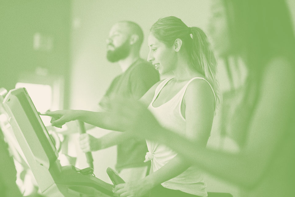 Three people use exercise machines at a gym in Briggs Ranch, with one woman in the foreground pressing buttons on a control panel—a glimpse of active living in this master-planned community.