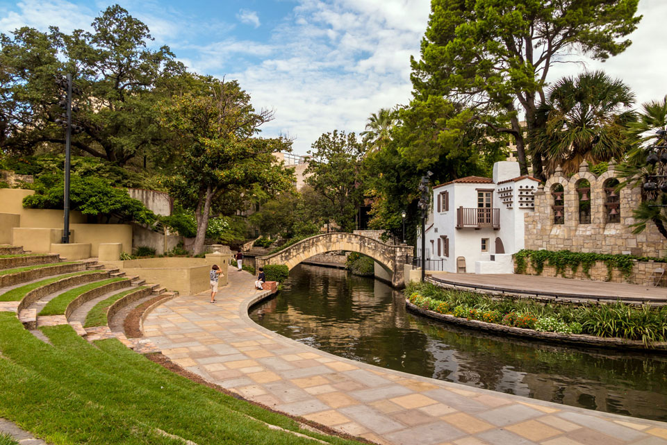 Stone amphitheater and walkway beside a canal in Medina Valley, with a small arched bridge and a white building surrounded by trees and greenery—an inviting spot near the new homes at Briggs Ranch.