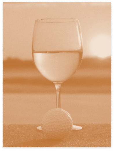 A wine glass filled with water stands behind a golf ball on a flat surface, set against the blurred backdrop of a master-planned community in Medina Valley at sunset.