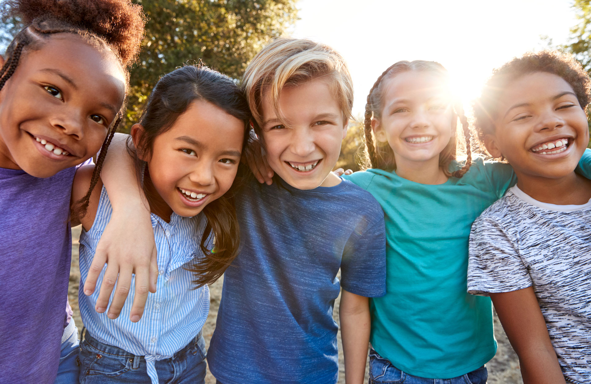 Five smiling children stand close together outdoors in a master-planned community, arms around each other, with sunlight shining through trees and new homes for sale in the background.