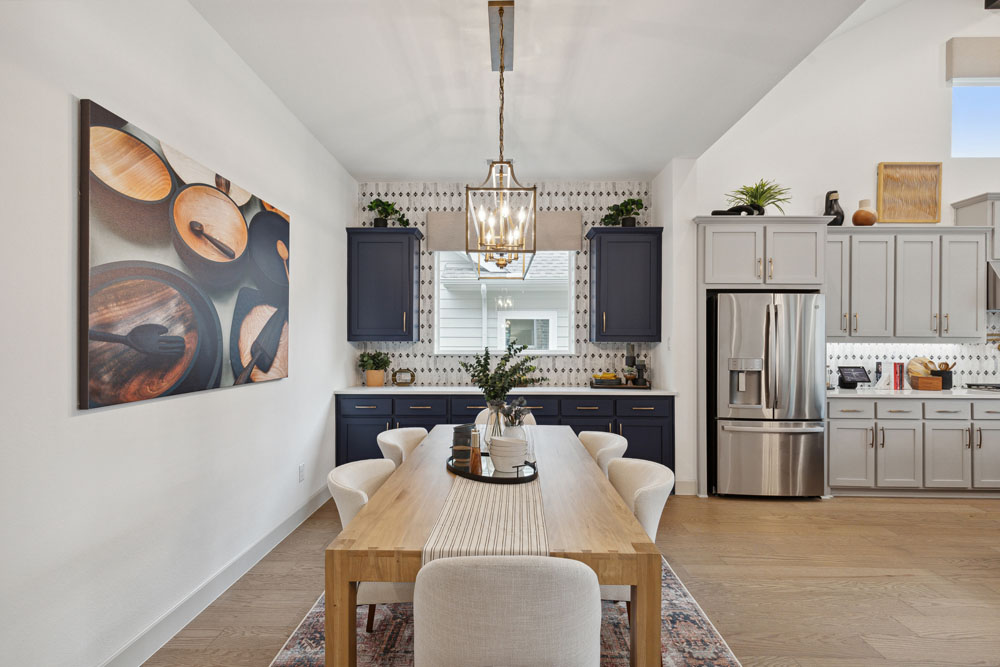 Modern dining area in Briggs Ranch features a wooden table, six upholstered chairs, blue cabinetry, stainless steel appliances, and a large kitchen-themed artwork—perfect for gatherings in these stylish new homes near Medina Valley.