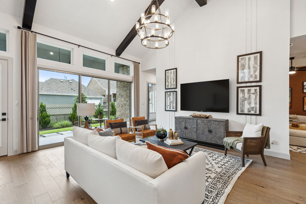 Modern living room with a white sofa, armchairs, large TV, decorative rug, chandelier, and spacious windows overlooking a backyard in the master-planned community of Briggs Ranch near San Antonio.