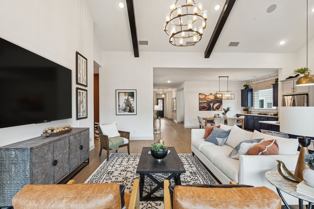 Modern open-concept living room with a white sofa, leather chairs, large TV, patterned rug, and chandelier, adjoining a kitchen and dining area with wood floors and exposed ceiling beams—perfect for new homes in the heart of Medina Valley.