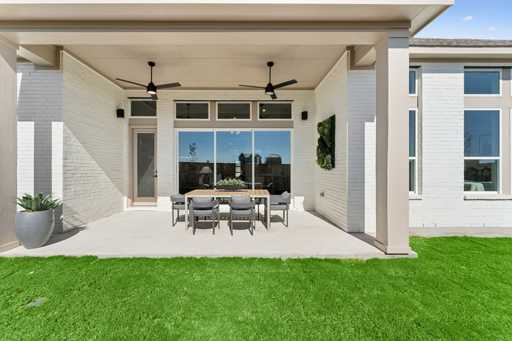 Covered patio with dining table, six chairs, ceiling fans, and green artificial grass lawn.