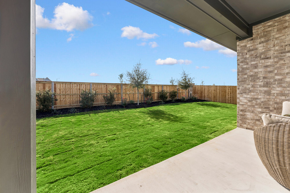 View of a fenced backyard with green grass and small trees, seen from a covered patio.