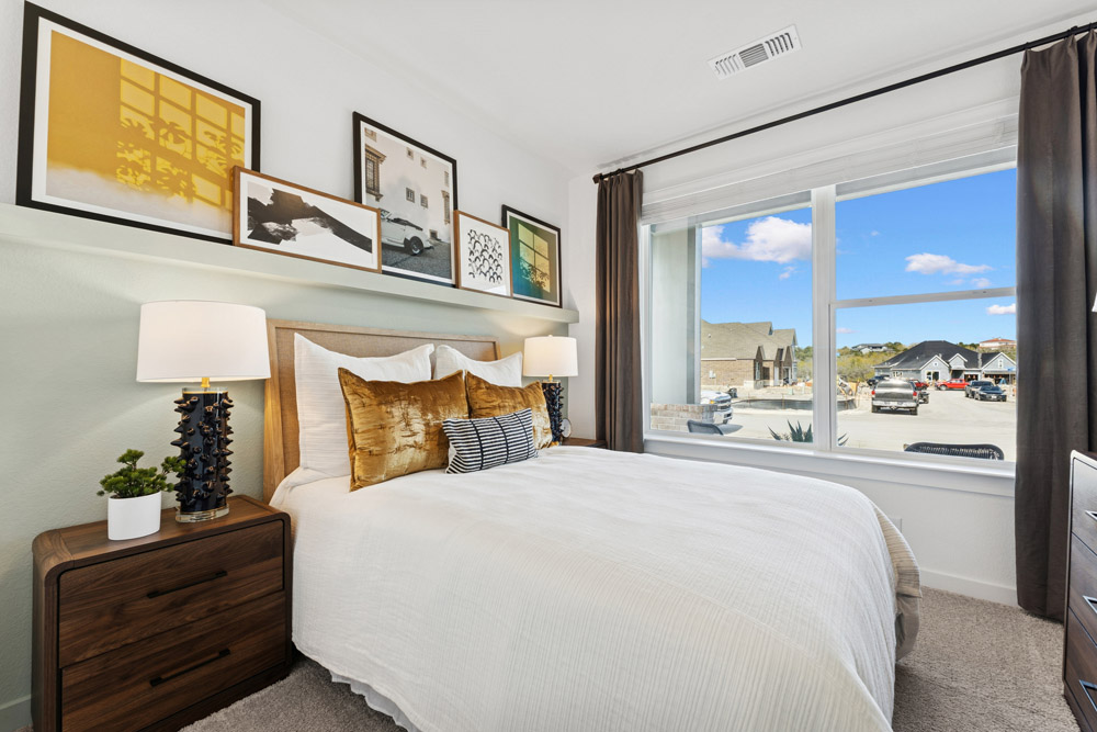 A neatly made bed with gold pillows beside a window showing a suburban street view.
