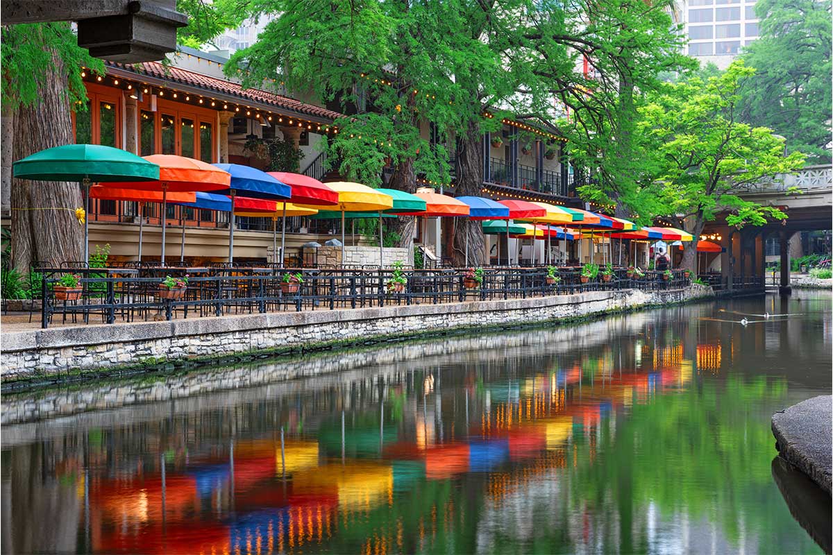 Outdoor café with colorful umbrellas lines a riverside walkway, reflected in the calm water.