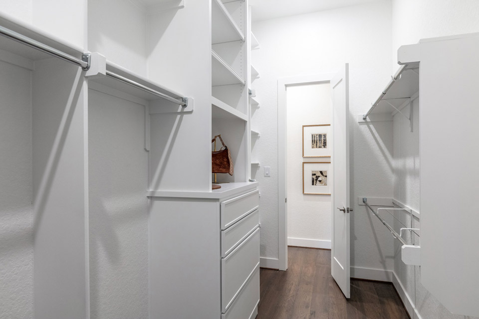 Walk-in closet with white shelves, hanging rods, wood floor, and an open door to hallway.