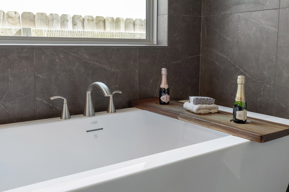 Modern bathtub with a wooden tray holding towels and two bottles of sparkling wine.