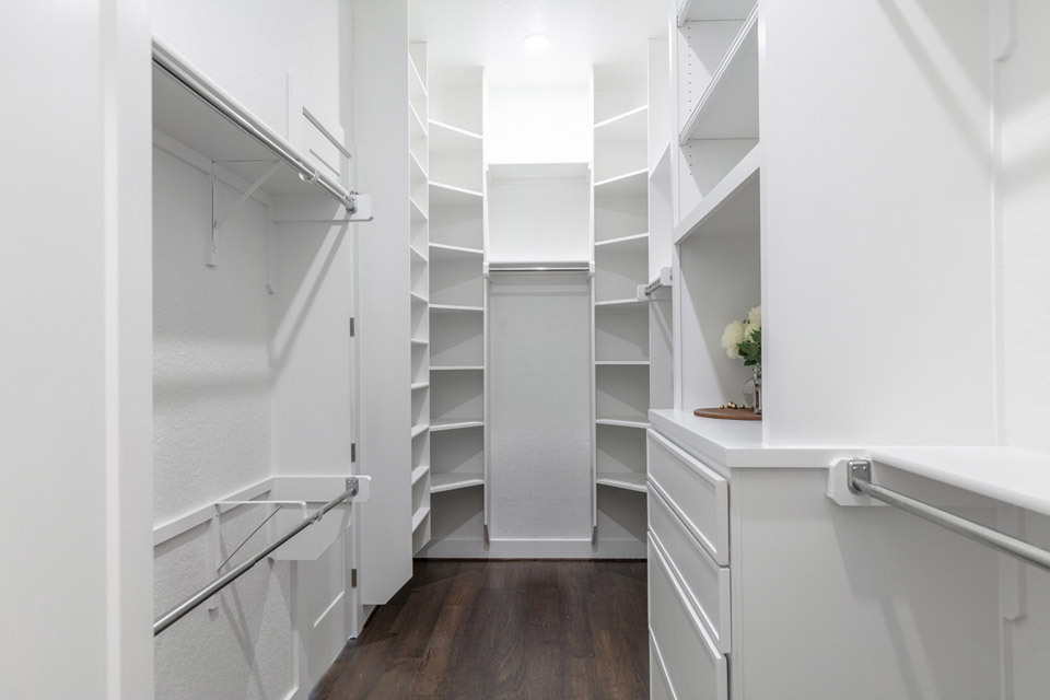 Walk-in closet with white shelves, drawers, hanging rods, and dark wood flooring.