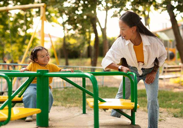 Woman spinning a little girl on a yellow merry-go-round at a playground on a sunny day.