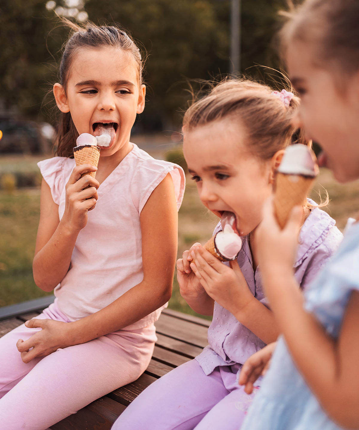 Three young girls sitting outside eating ice cream cones on a wooden bench.