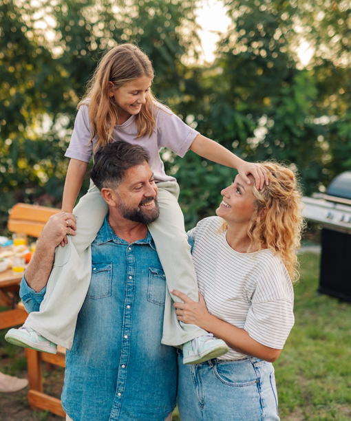 A man carries a girl on his shoulders while a woman smiles at them outdoors near a grill.