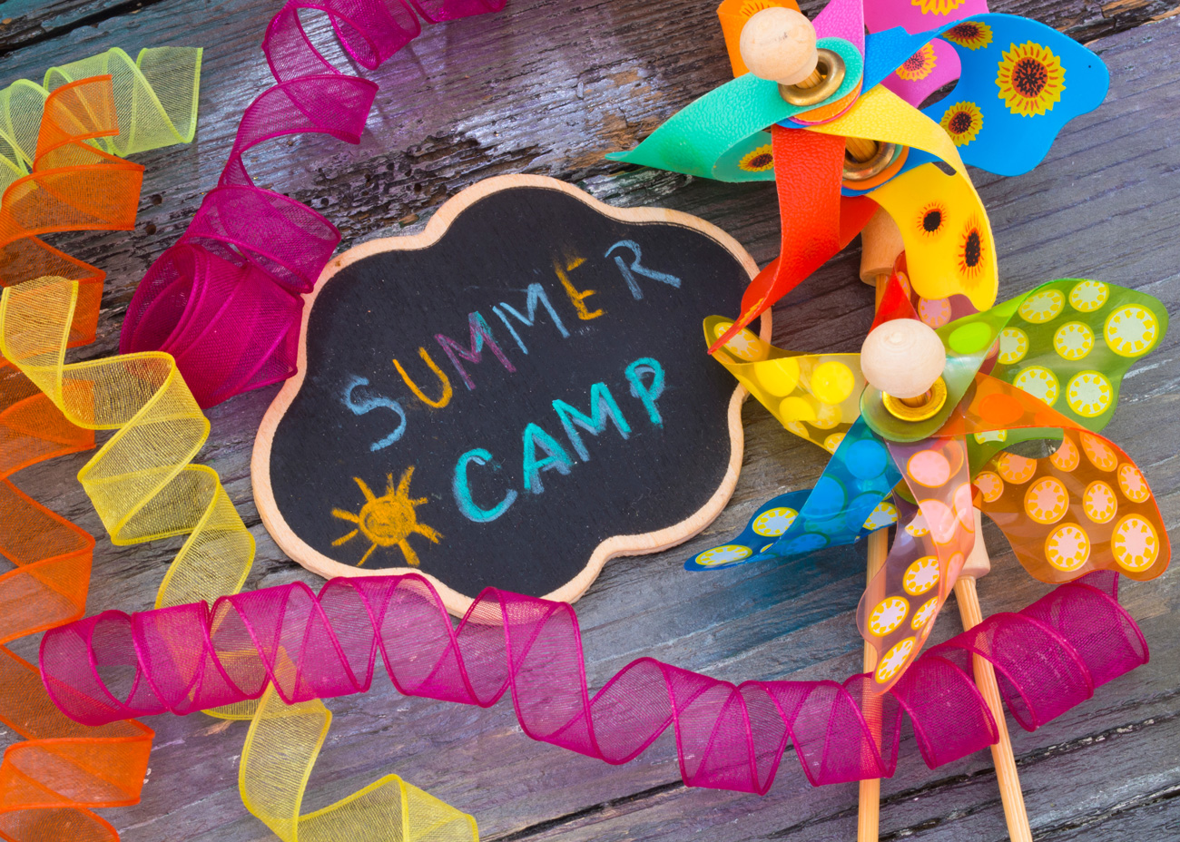 Chalkboard sign reading Summer Camp with colorful ribbon and pinwheels on wooden surface.